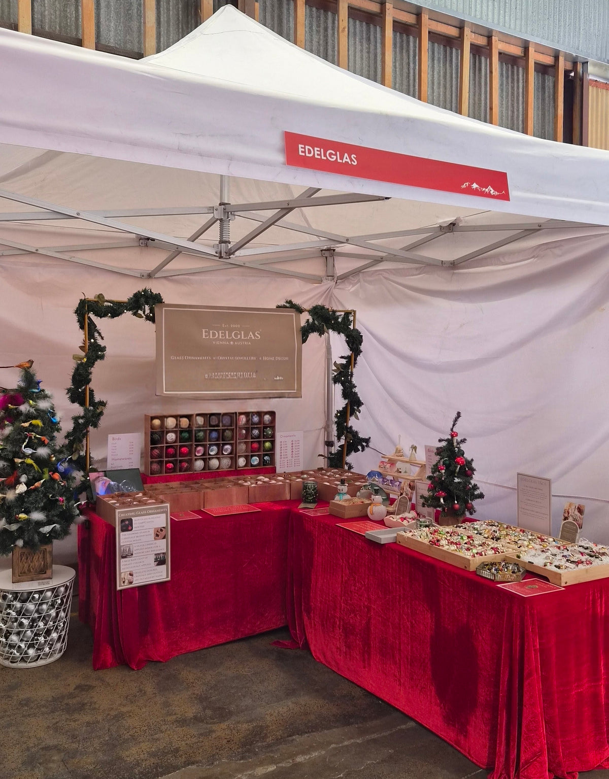 An outdoor market booth under a white canopy tent branded EDELGLAS, with red velvet tables, holiday garlands, small decorated trees, and assorted packaged goods displayed on the tables.